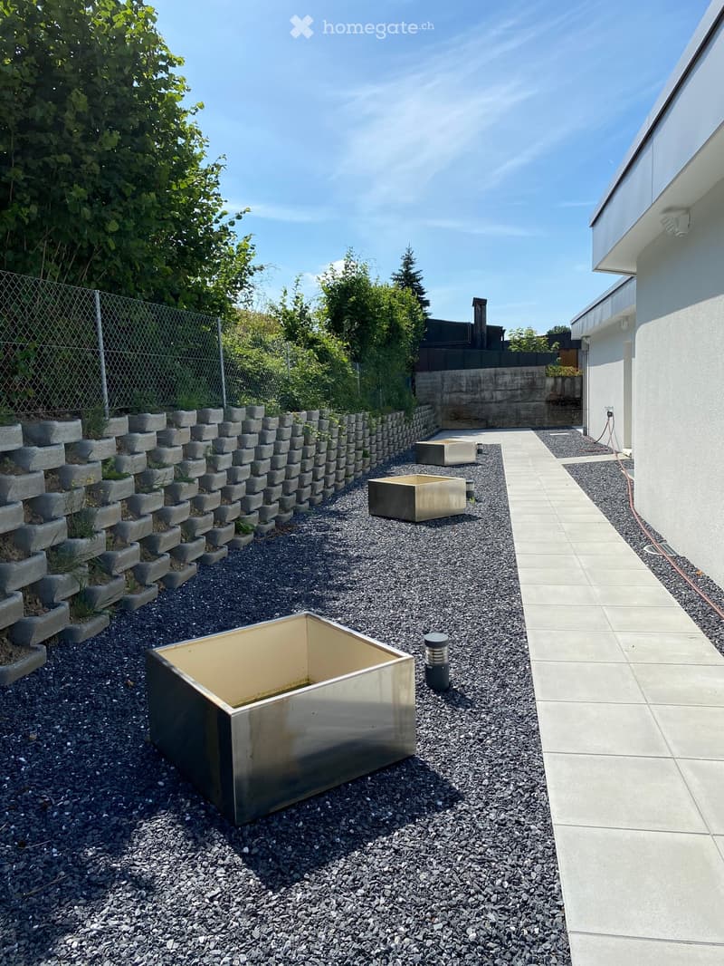 Modern garden area with gravel ground, raised concrete planters, and a metal planter box. Features a paved pathway and a small fence.