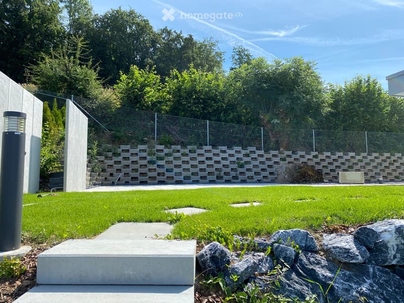 Well-maintained garden with a raised concrete retaining wall, neatly trimmed grass, decorative rocks, and a modern outdoor light fixture.