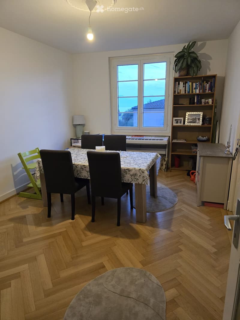 Dining area with wooden herringbone flooring, a round table with four black chairs, a piano, a bookshelf, and a small green chair.