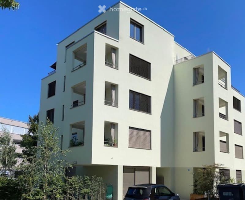 Modern multi-story residential building with light-colored facade, multiple windows, balconies, and a parking area at the base.