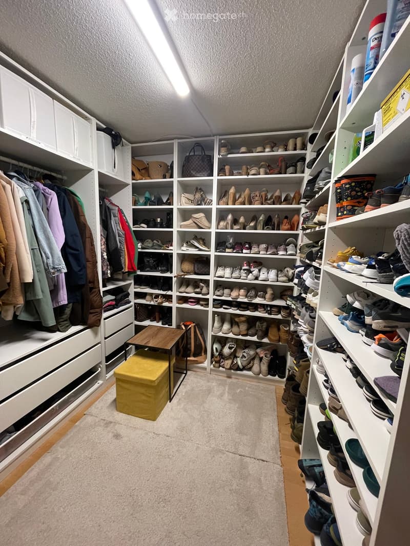 Well-organized walk-in closet with white shelving units filled with shoes, handbags, and clothing. A small wooden table and yellow stool are placed in the center.