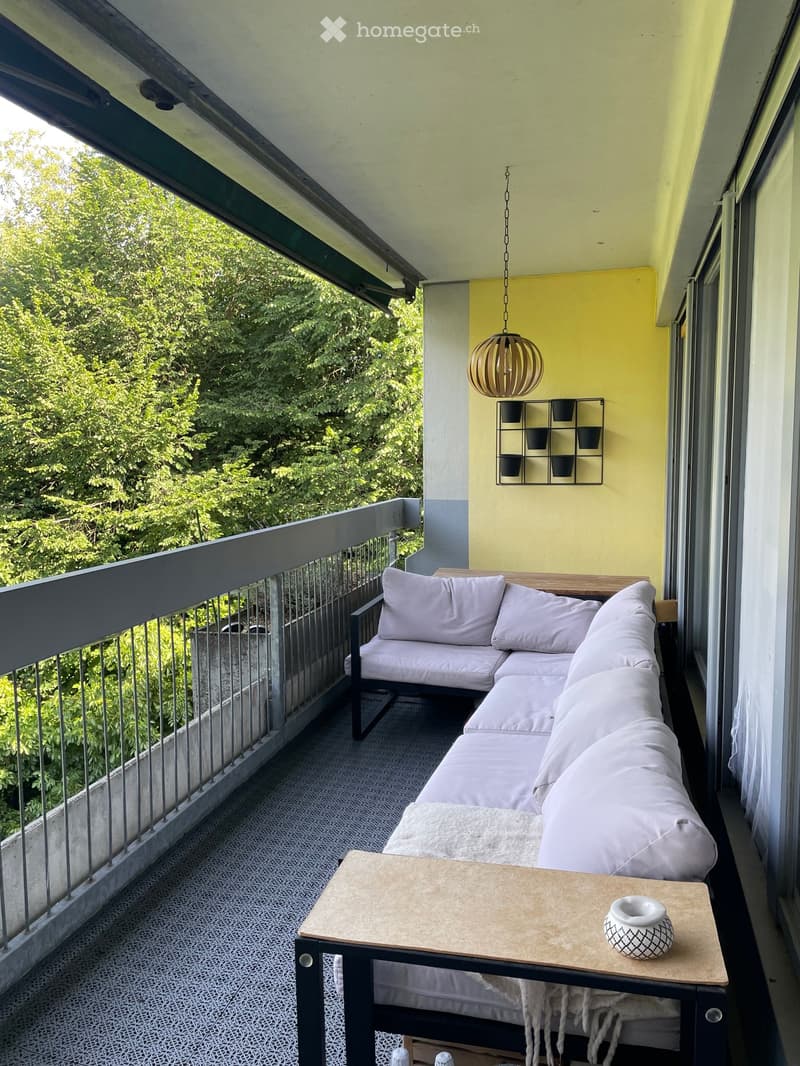 Balcony with a grey sectional sofa, small wooden table, and decorative vase, overlooking greenery.