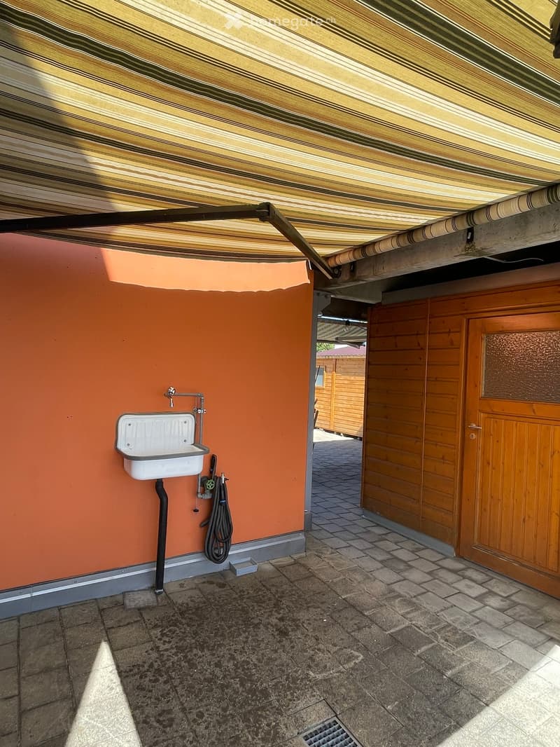 Outdoor area with a covered roof, tiled floor, and a white sink mounted on an orange wall. A wooden door leads to another area, and a hose is hanging nearby.