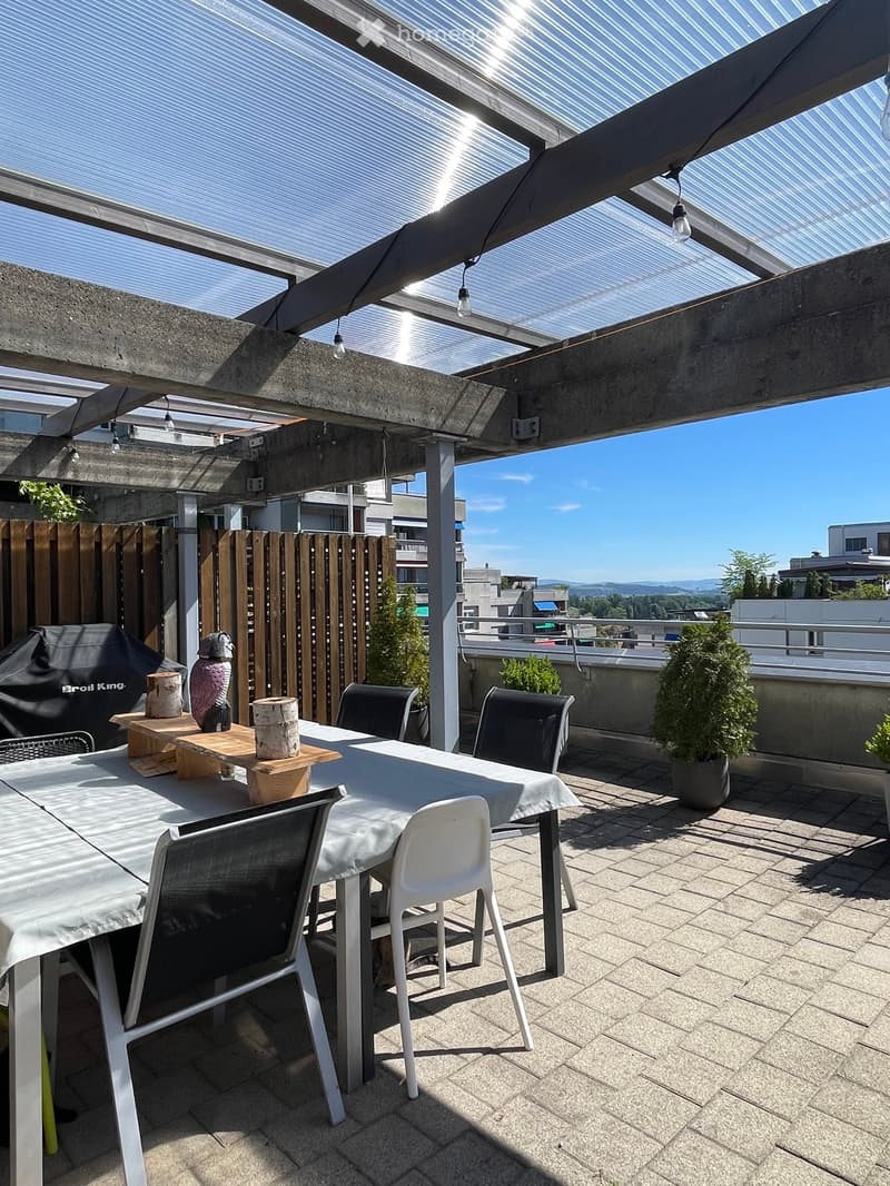 A modern rooftop terrace with a dining table, chairs, an umbrella, a grill, and potted plants, featuring a clear sky and cityscape view.