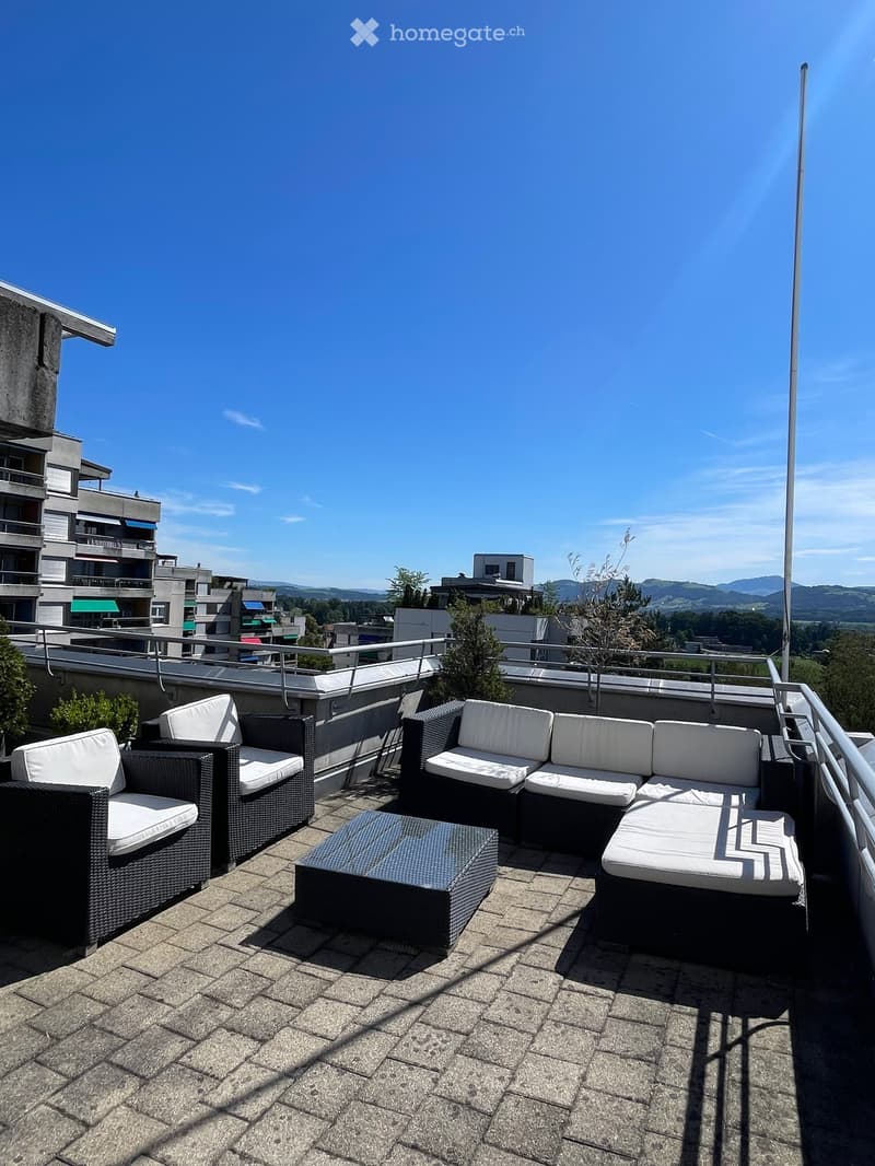 A modern rooftop terrace with a sofa, chairs, and a table, overlooking a cityscape with buildings and mountains in the background.
