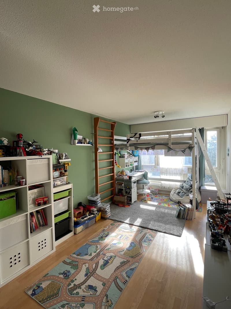 A well-lit children's bedroom with a bunk bed, desk, bookshelf, and various toys. The room features a colorful play mat and a cozy beanie chair.