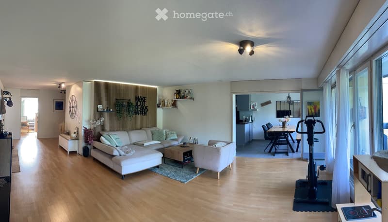 Modern living and dining area with light wood flooring, white sofa, coffee table, armchair, dining table with chairs, exercise bike, and kitchen area with cabinets and counter.