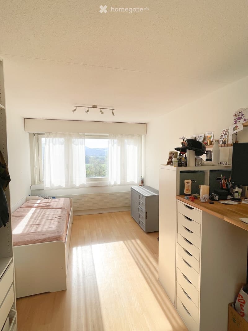 A small, well-lit bedroom with a single bed, a desk, and a window with sheer curtains. The room features light wooden flooring and white walls.