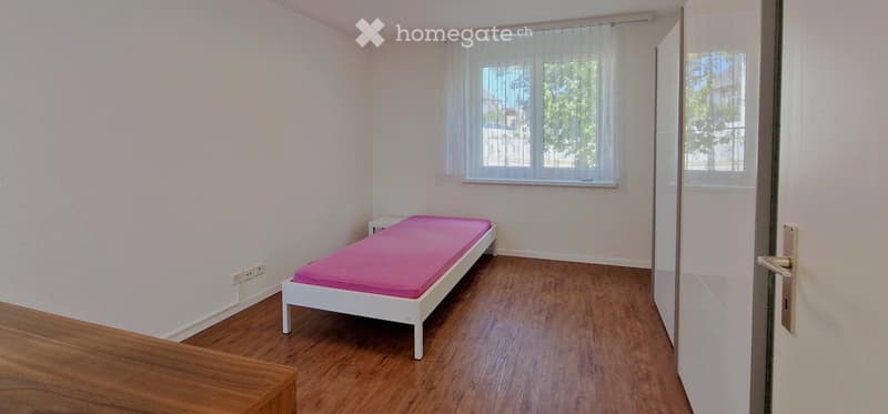 Small bedroom with a single bed, pink mattress, nightstand, wooden flooring, and large window with sheer curtains.