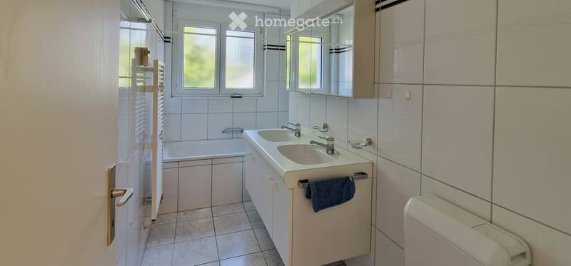 Modern bathroom with a bathtub, double sink, mirror cabinet, and a towel hanging on the side. The room features tiled walls and floor, with a window providing natural light.
