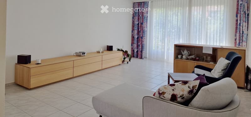 Bright living room with a grey sofa, wooden coffee table, armchair, and a wooden cabinet with a teapot and cups. The room features large windows with curtains and a tiled floor.