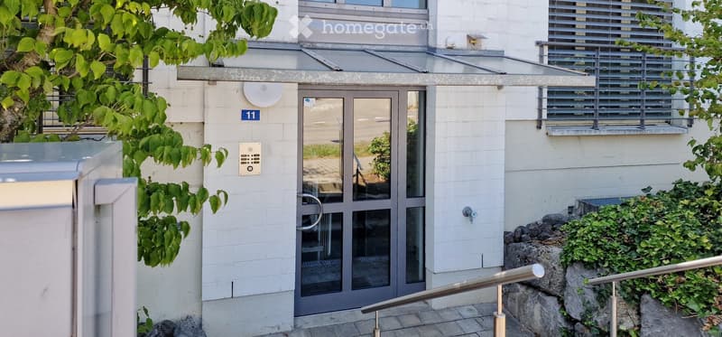 Entrance to a modern building with a glass door, intercom system, and greenery around the entrance.