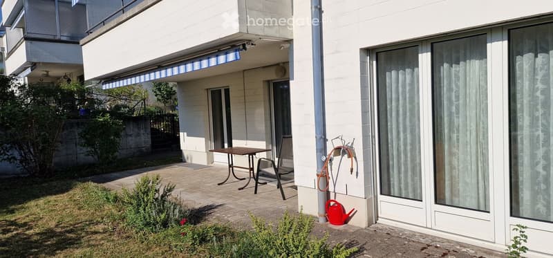 Balcony area with a table, chair, garden hose, and watering can, adjacent to a building with large windows and a retractable awning.