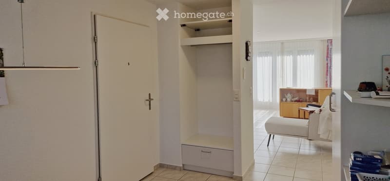 A modern, well-lit room with a white sofa, small table, and built-in storage. The space features large windows with sheer curtains and a minimalist design.