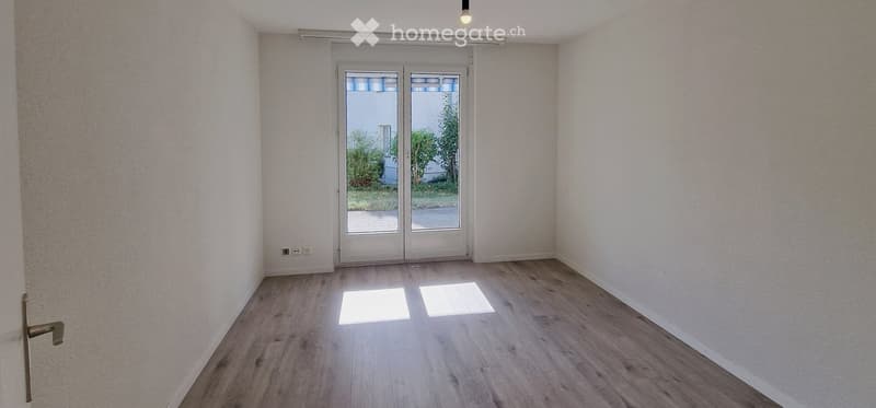 Empty room with light wooden flooring, white walls, and a large glass sliding door leading outside.