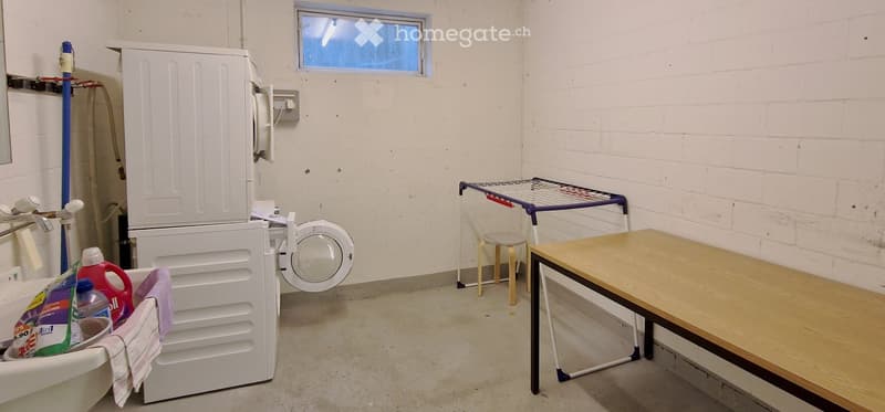 Laundry room with a washing machine, drying rack, small stool, wooden table, and detergent bottles on a shelf.