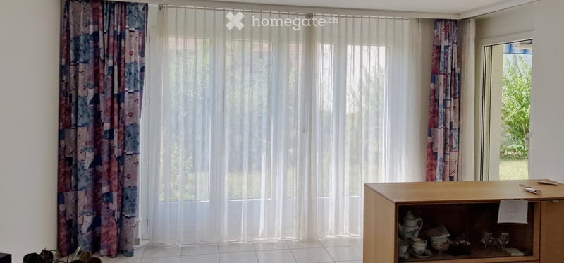 Bright room with large windows covered by sheer white curtains and colorful side curtains. A wooden display cabinet holds various decorative items including a teapot, cups, bowls, and glasses.