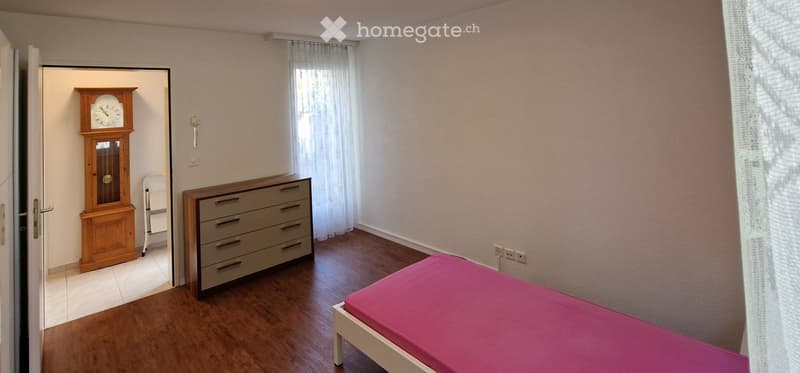 Bedroom with wooden floor, pink bedspread, chest of drawers, grandfather clock, and a chair in the corner.
