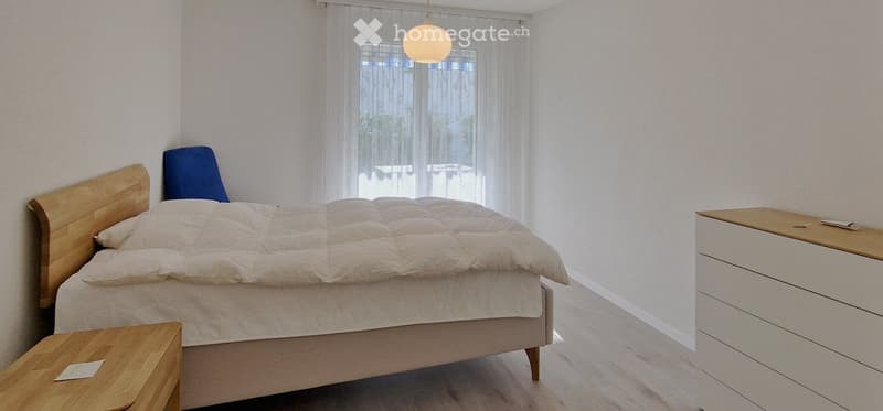 Modern bedroom with a wooden bed, white bedding, a nightstand, and a drawer unit. Natural light filters through sheer curtains.