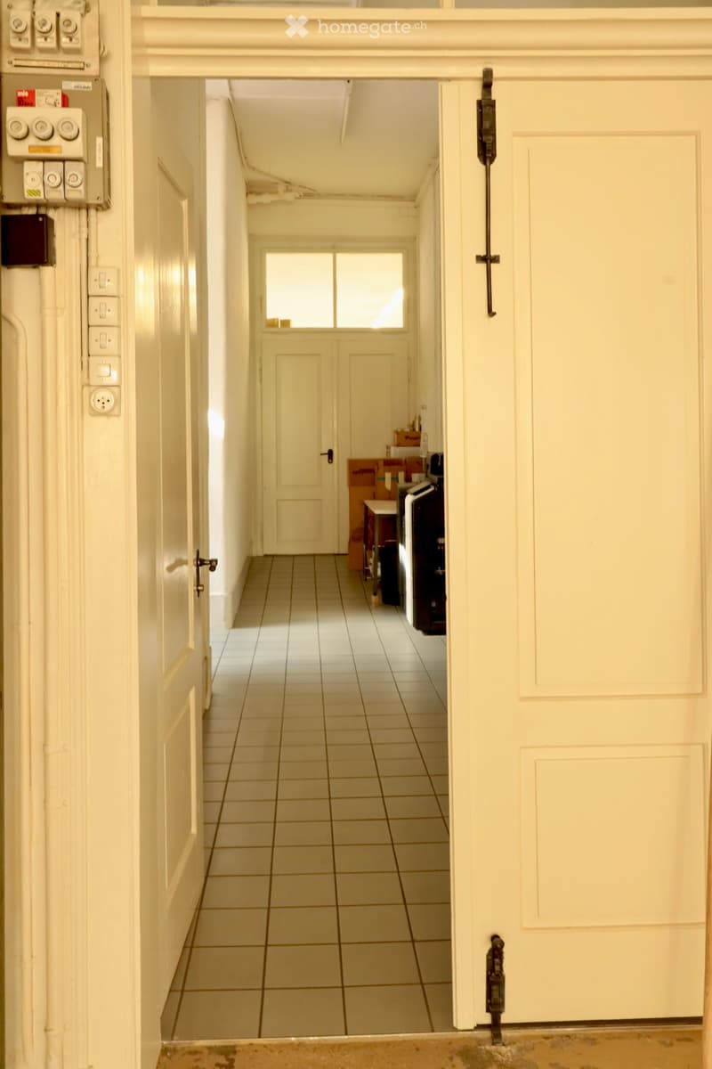 A narrow hallway with tiled flooring, white walls, and several doors leading to other rooms. A window at the end provides natural light, and electrical panels are visible on the left wall.