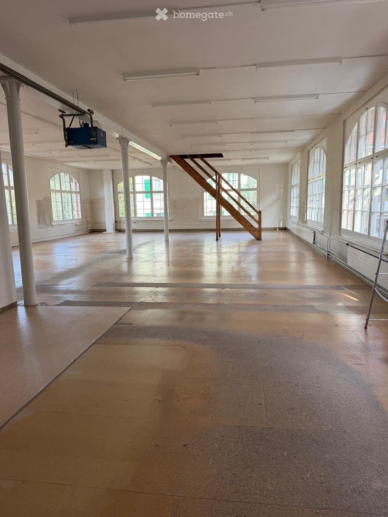 Large, empty room with wooden flooring, white walls, large windows, and a wooden staircase leading to an upper level. Exposed beams and columns are visible.