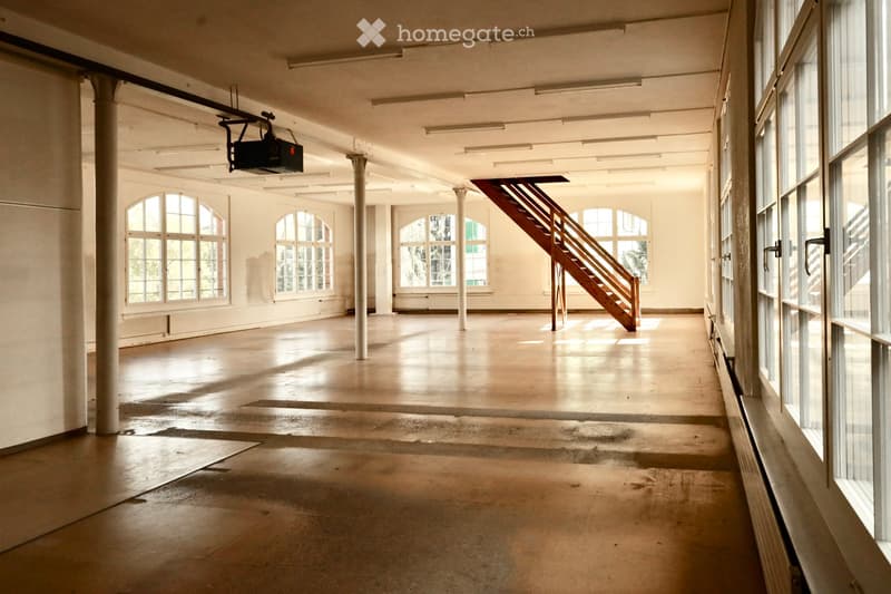 Empty industrial-style room with large windows, concrete floor, exposed metal columns, and a wooden staircase leading to an upper level.