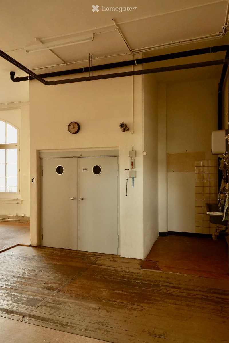 Empty room with wooden flooring, white walls, a large window, a closed door, and a clock on the wall. No furniture or equipment is present.