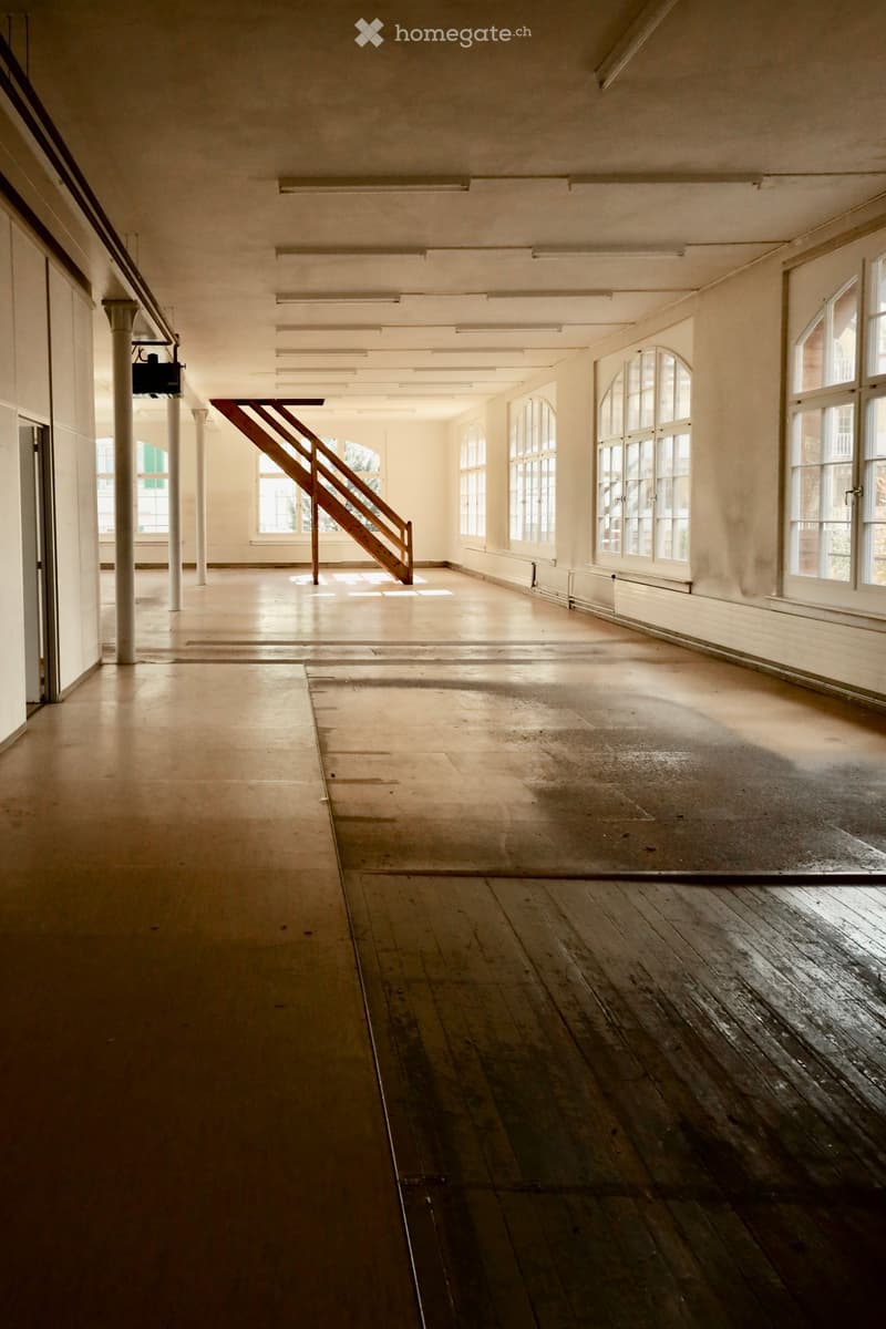 Empty room with wooden floors, large windows, and a staircase leading to another level. The space is well-lit with natural light and has exposed structural elements.