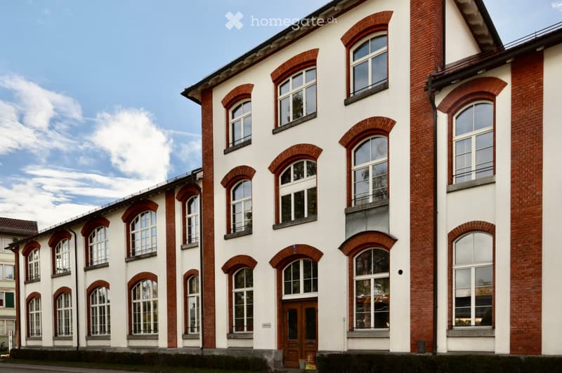 A multi-story building with white walls and red brick accents, featuring numerous large windows with arched frames and a central wooden door.