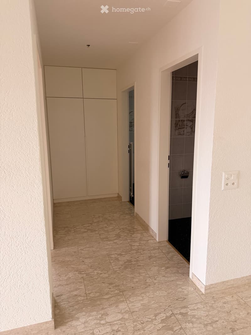 Empty hallway with beige tiled floor, white walls, built-in closet, and a partially visible bathroom with dark tiles and a glass door.