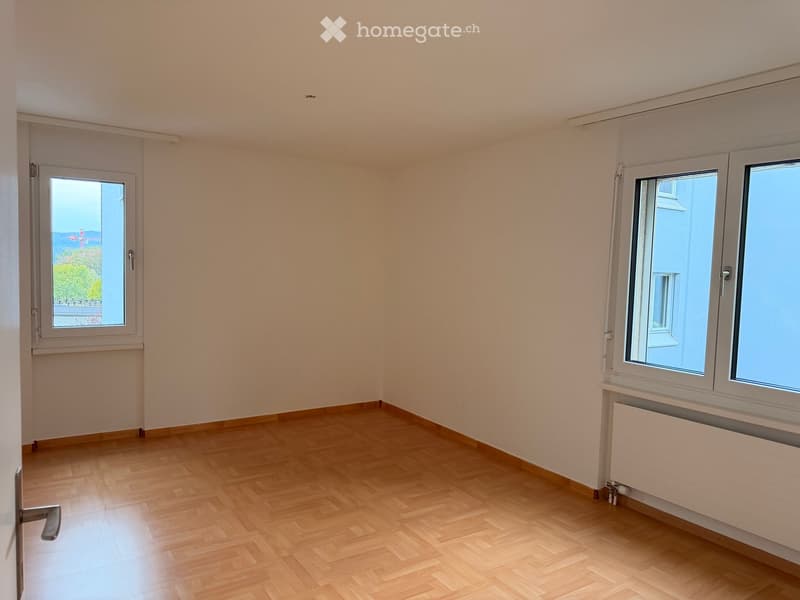 Empty room with light-colored walls, wooden flooring, and two windows with white frames. Radiators are visible beneath the windows.