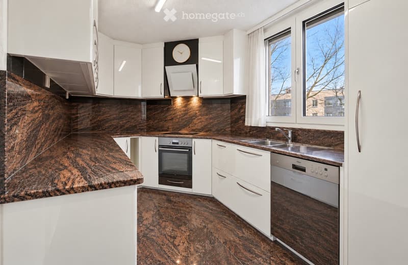 Modern kitchen with white cabinets, dark granite countertops, and a stainless steel oven and dishwasher. Features a large window with white curtains and a clock on the wall.