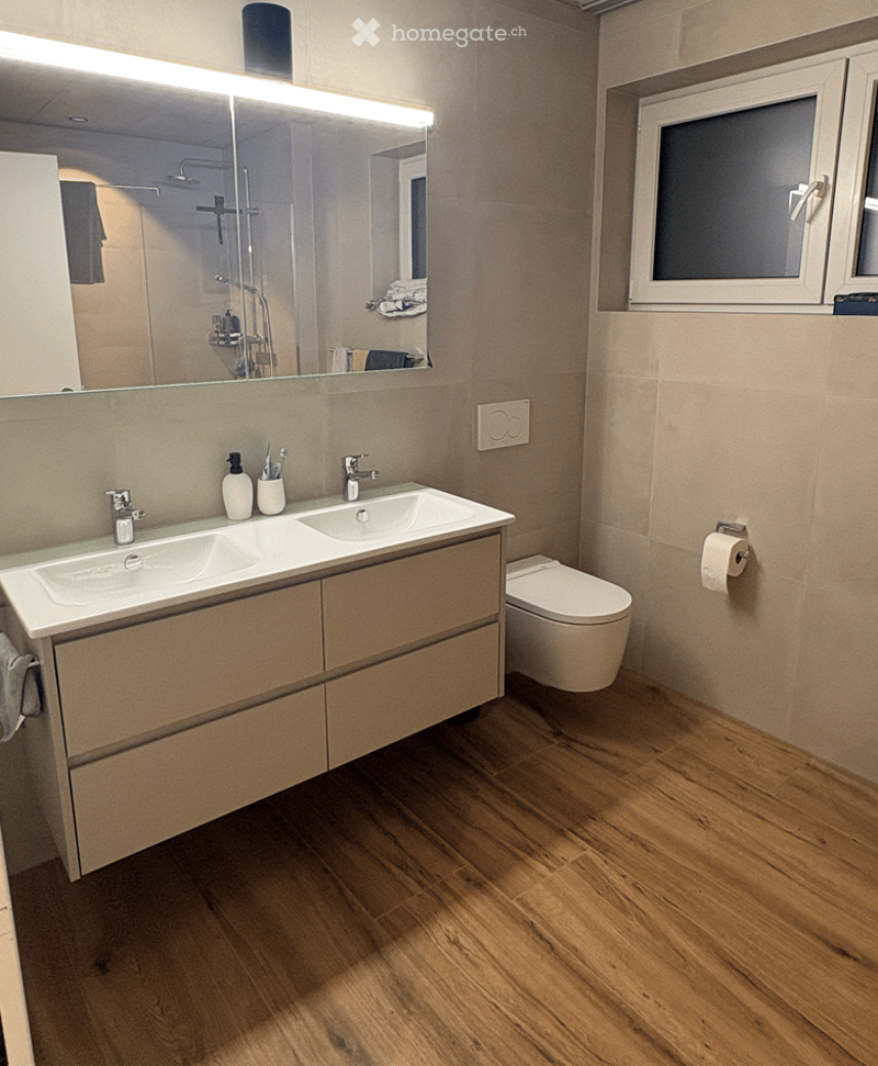 Modern bathroom with double sink, large mirror, toilet, and wooden flooring.