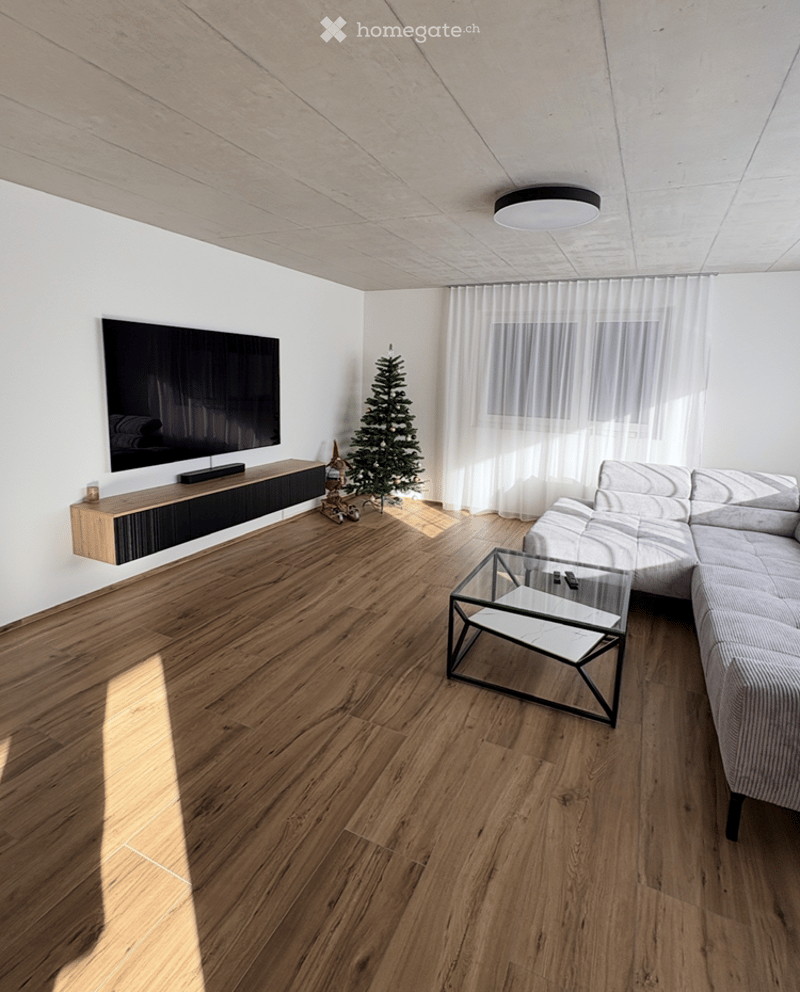 Modern living room with light wooden flooring, white walls, a large sectional sofa, a glass coffee table, a flat-screen TV on a wooden stand, and a small Christmas tree near the window with sheer curtains.