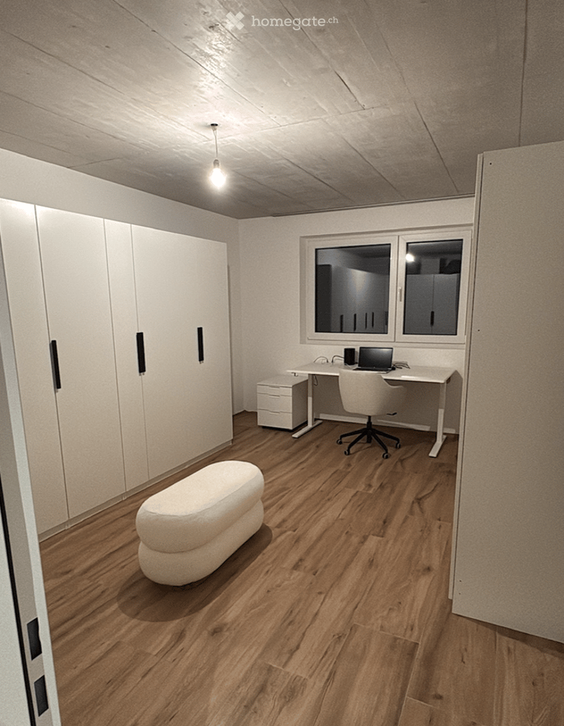 A modern, minimalist room with light wooden flooring, white walls, and a sleek white desk setup. Features include a white ergonomic chair, a small white stool, a laptop on the desk, and built-in white cabinets with black handles. The room is well-lit with