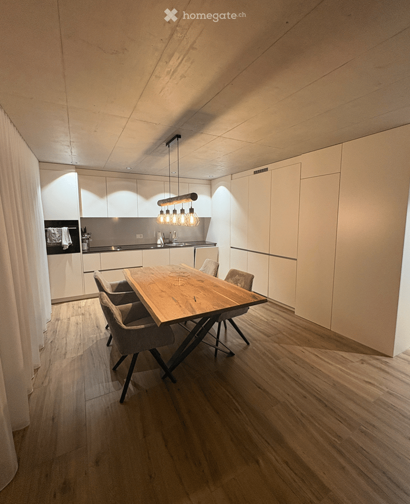 Modern kitchen with white cabinets, a wooden dining table, four chairs, and a hanging light fixture above the table. The floor is made of light wood, and the backsplash is sleek and contemporary.
