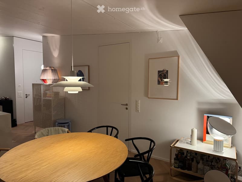 A modern, well-lit room with a round wooden table, black chairs, a small shelf with bottles and a lamp, and a hanging pendant light. The room features white walls, wooden flooring, and a large window allowing natural light to stream in.