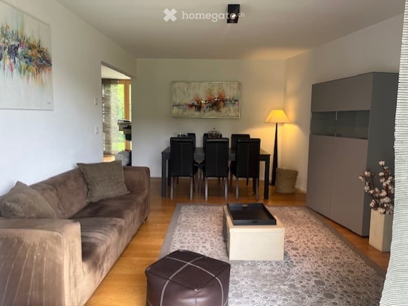 Modern living and dining area with a brown sofa, ottoman, coffee table, dining table with chairs, floor lamp, and cabinet. Artwork adorns the walls, and a vase with flowers sits on the floor.