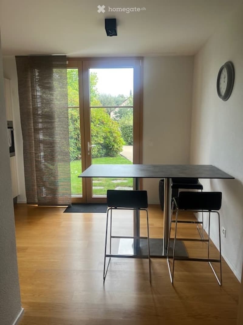 Modern dining area with a sleek black table and two matching stools, large glass door leading to a garden, wooden flooring, and a curtain on the window.
