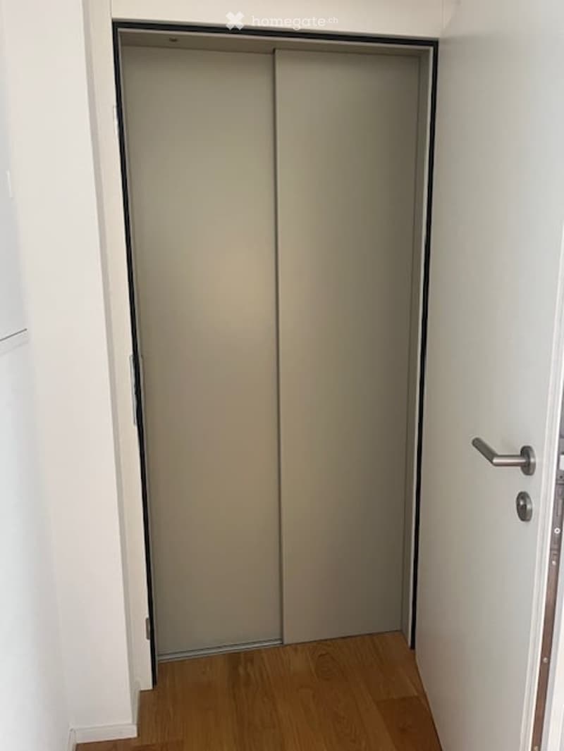 A narrow hallway with light wooden flooring and white walls, featuring a closed sliding door on one side and a standard door on the other.