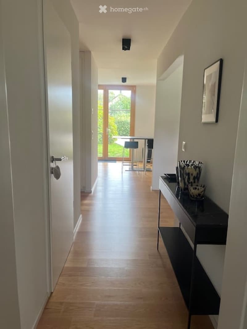 A modern hallway with light wooden flooring, white walls, and a sleek black console table with decorative items. A glass door at the end leads to a bright room with a bar table and chairs.