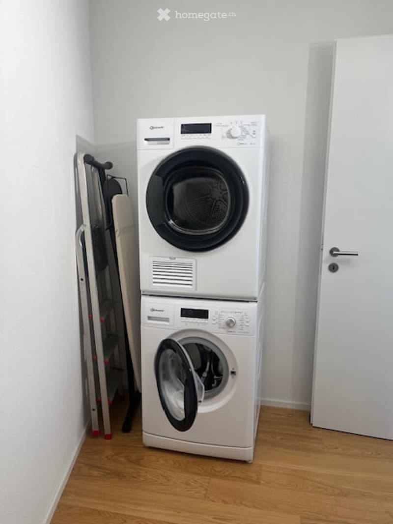 A small room with a white washing machine and dryer stacked vertically, a wooden floor, and a white door.