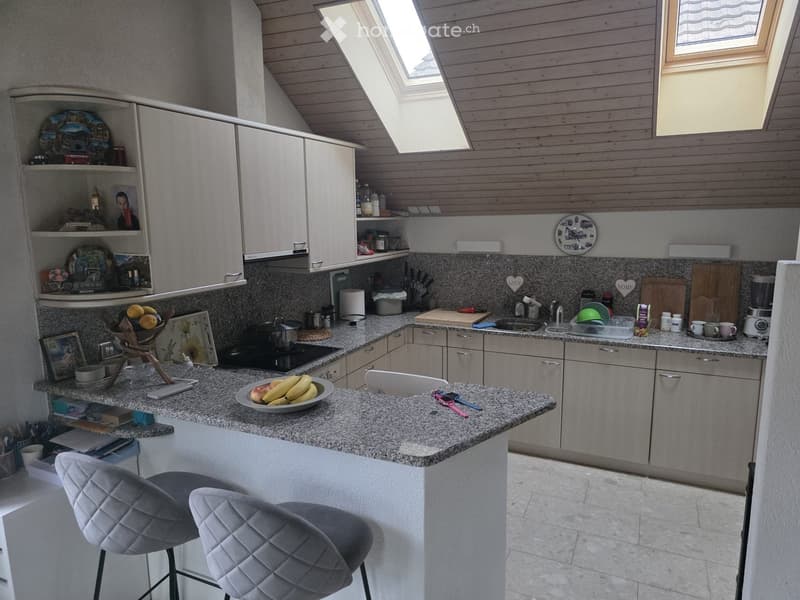 Modern kitchen with light grey cabinets, granite countertops, and a central island with two grey upholstered stools. The kitchen features a sink, stove, and various appliances, with decorative items and fruit on the counter.