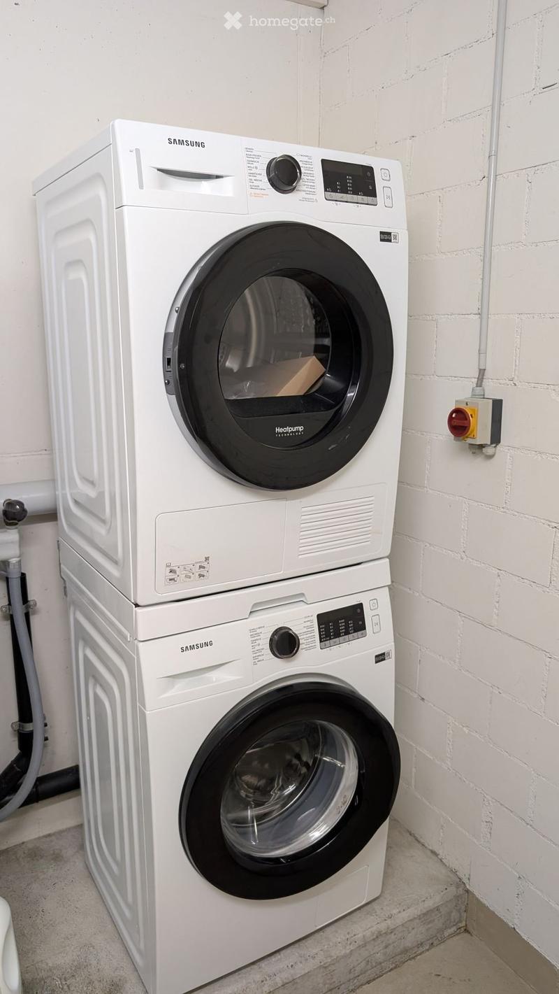 Stacked washing machine and dryer, both Samsung, white in color, digital control panels, placed against a brick wall