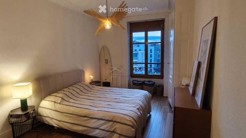 Cozy bedroom with a large bed, nightstand with lamp, picture frame on dresser, surfboard leaning against wall, and a view of the city through the window.