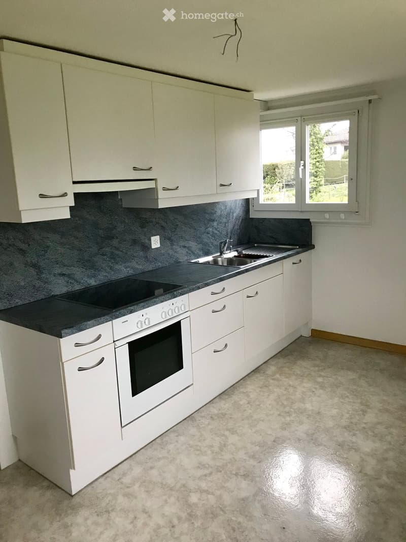 Modern kitchen with white cabinets, a sink, an oven, and a dark countertop. The walls are painted in a dark gray color, and the floor is covered with a light gray, polished concrete.