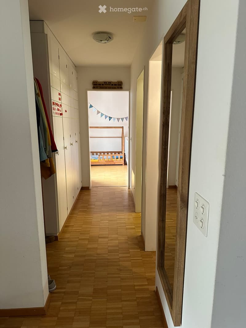 A long hallway with wooden flooring, white walls, and built-in white cabinets on one side. A mirror is mounted on the right wall, and a small decorative banner is visible in the background.