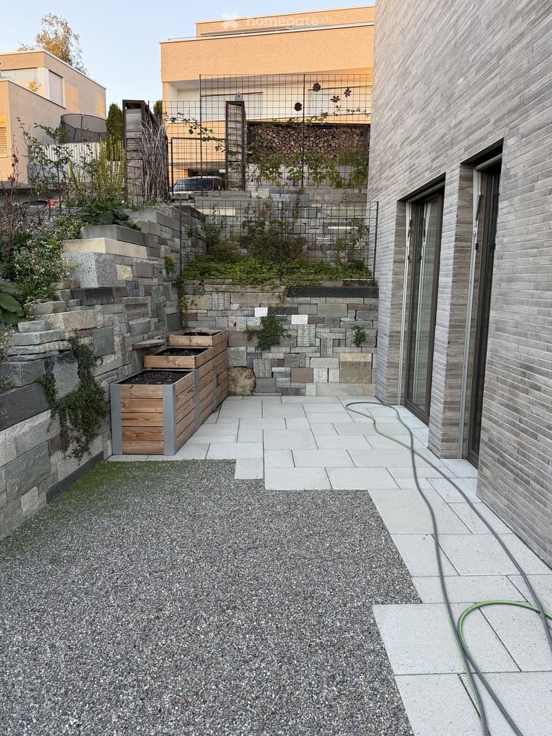 Modern outdoor space with stone and gravel surfaces, wooden bench, greenery, and a raised garden bed.