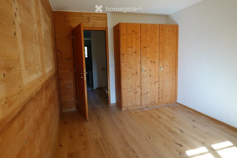 Empty room with wooden flooring, white walls, and a large wooden wardrobe. An open doorway leads to another room.