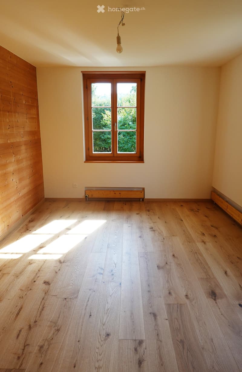 Empty room with wooden flooring, light-colored walls, and a large window with wooden frame allowing natural light to enter.